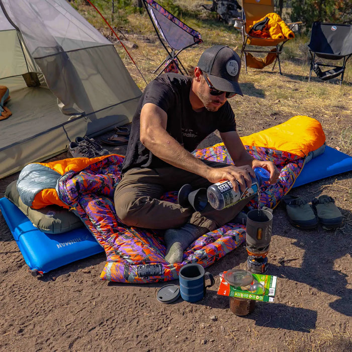 A man in a cap sits on the Big Agnes Anthracite 20° - Alpenglow recycled synthetic mummy sleeping bag and an inflatable pad beside a tent, pouring water into a stove for coffee while camping; mugs, camping gear, and chair are nearby.
