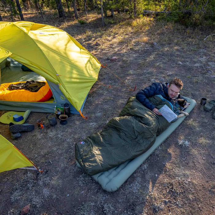 A man relaxes in a Big Agnes Anthracite 30° mummy sleeping bag on a camping mat by a yellow tent, reading outdoors. Camping gear, boots, and a mug are scattered nearby as sunlight filters through the trees.