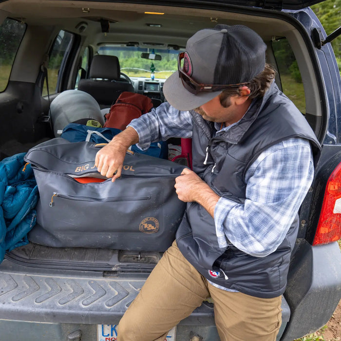 Wearing a cap and vest, a person unzips the Big Agnes Big Joe Gear Duffel on an SUV's open tailgate, surrounded by outdoor equipment like a blue sleeping bag and assorted bags.