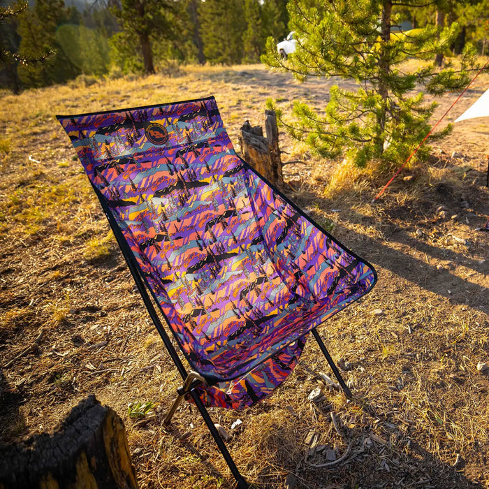 The Big Agnes Big Six Camp Chair - Alpenglow, featuring a colorful, patterned design, is set up on dry grass near pine trees. Sunlight casts warm shadows on the chair and ground, creating an inviting outdoor atmosphere.