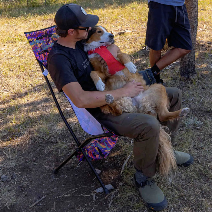 Seated outdoors on dry grass in a Big Agnes Big Six Camp Chair - Alpenglow, a person cradles and kisses their relaxed dog, while another stands nearby amid scattered trees.