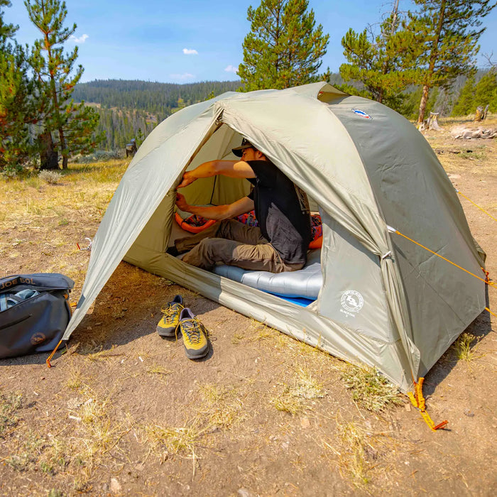 A person sits inside the Big Agnes Blacktail 2 - Vetiver tent set up in a forest clearing, organizing gear. Hiking shoes and a backpack rest outside the tent, with evergreen trees and distant hills in the background.