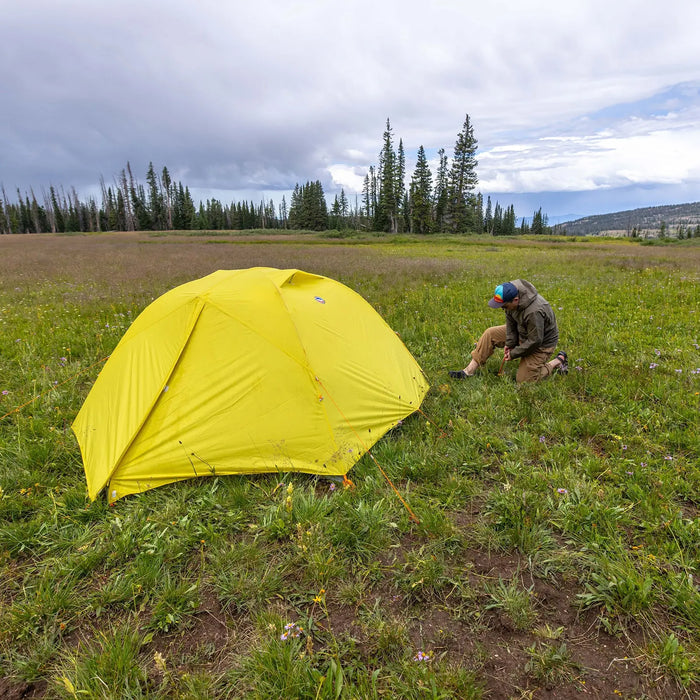 A person kneels beside a Big Agnes Blacktail 2 tent set up in a grassy field among pine trees, with distant hills and a cloudy sky overhead.