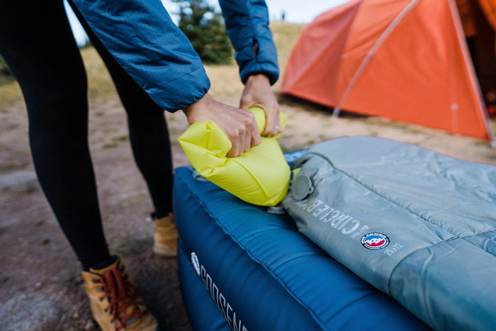 A camper inflates a yellow Circle Back™ Insulated sleeping pad by Big Agnes beside a blue and gray camping pad outside near an orange tent.