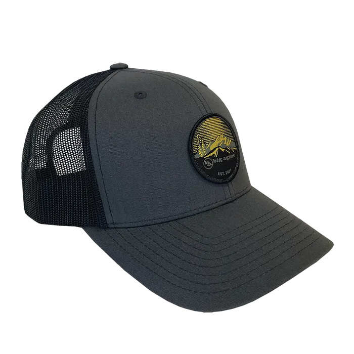 The Big Agnes EST. 2001 Logo Baseball Hat is a dark gray and black trucker hat with a round front patch of a yellow mountain scene, curved bill, adjustable mesh back, and celebrates Big Agnes' 25th Anniversary.