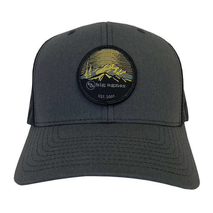 The EST. 2001 Logo Baseball Hat by Big Agnes is a dark gray trucker cap with mesh sides and a structured brim, featuring a round patch with yellow mountains, trees, and “big agnes EST. 2001” on the front.