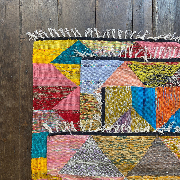 A colorful geometric patchwork rug with white fringe, laid flat on a dark rustic wooden plank floor.
