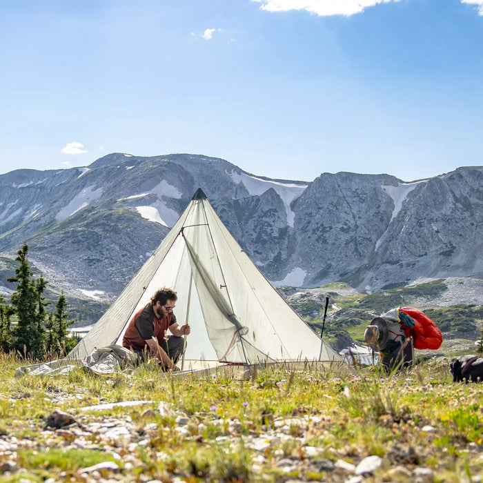 A camper relaxes beside a triangular tent featuring the Big Agnes Gold Camp 3 Mesh Inner for bug protection, set in a scenic mountain landscape with rocky peaks, snowy patches, and camping gear like a red backpack nearby.