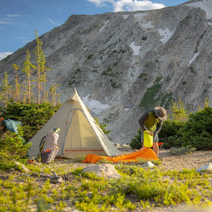 A camper sets up the Big Agnes Gold Camp 3 Tarp at a mountain campsite, surrounded by green shrubs and trees, with steep rocky cliffs rising in the background beneath a blue sky with scattered clouds.