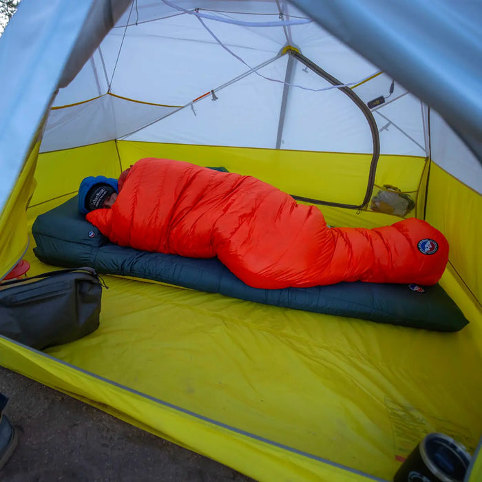 A person rests inside a yellow and white tent on a sleeping pad, wrapped in the Big Agnes Greystone 30° bright orange mummy-style sleeping bag, with camping gear visible nearby.