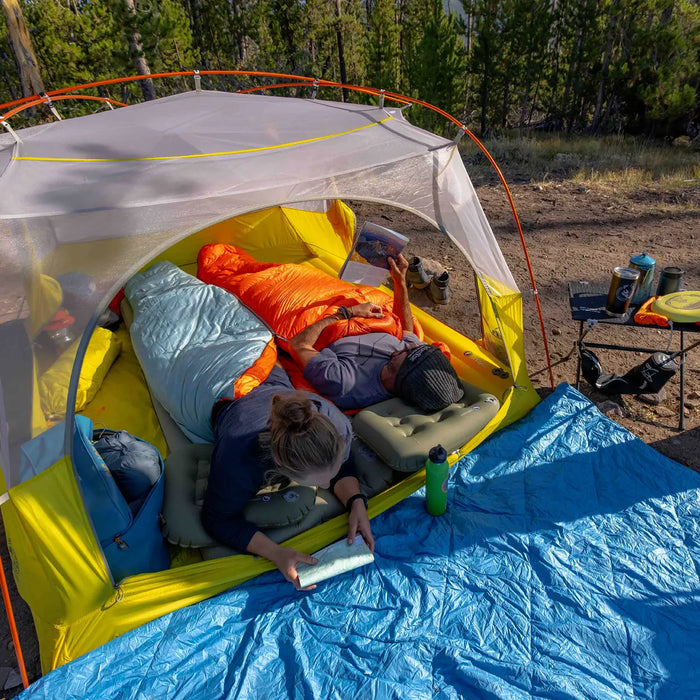 Two people relax in Big Agnes Greystone 30° mummy sleeping bags inside a tent pitched outdoors, one reading. Camping gear, shoes, and a water bottle are nearby on a blue tarp with trees in the background.