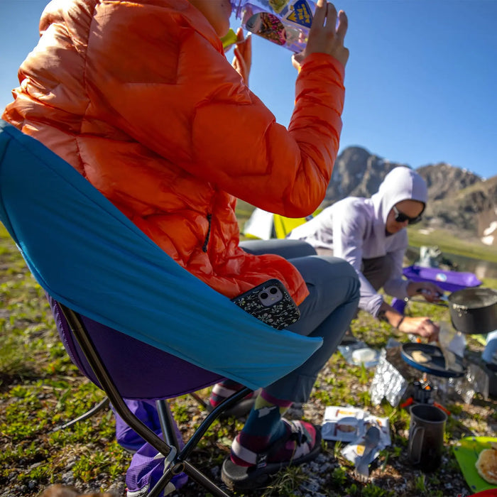 A person in an orange jacket relaxes on a Big Agnes Insulated Cover - Skyline UL Camp Chair, sipping a warm drink, while another cooks at a camp stove. Camping gear is on the grass, framed by mountains in the background.