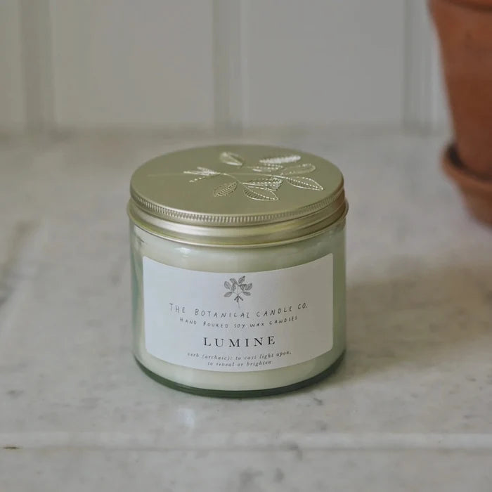A glass jar candle with a metallic gold lid and a white label reading 'The Botanical Candle Co. Lumine' sits on a light marble surface with a blurred potted plant in the background.