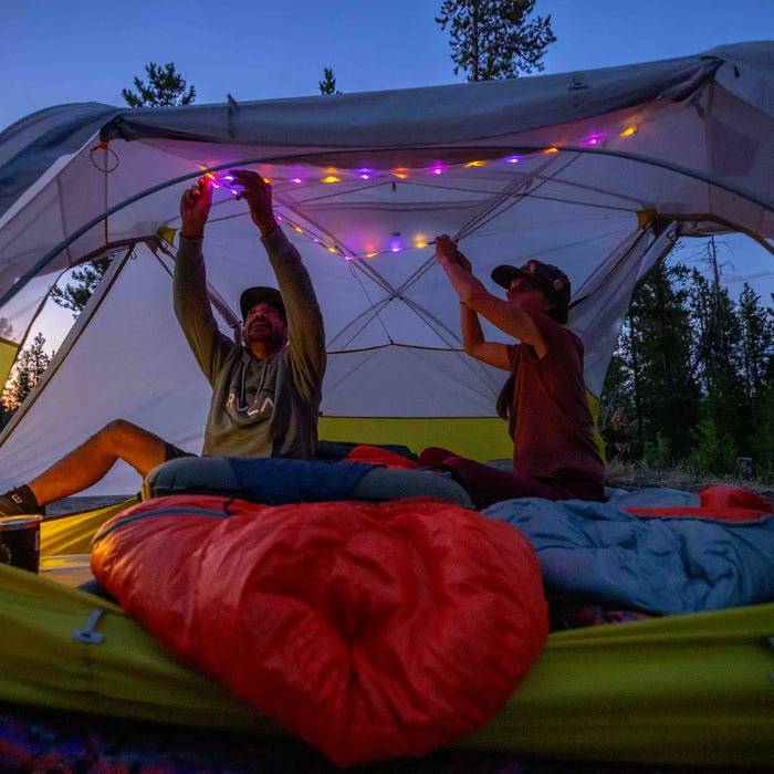 Two people sit in a tent at dusk, attaching the Big Agnes mtnGLO® Tent & Camp Lights - Alpenglow to the ceiling. Sleeping bags and gear are scattered nearby, with trees and a darkening sky creating a cozy campsite atmosphere.