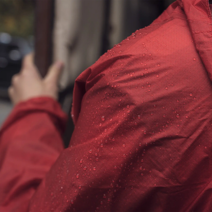 Wearing a red rain jacket treated with Nikwax TX Direct Spray-On Waterproofing by Big Agnes, a person stands outside holding a beige umbrella. The water-repellent outerwear and blurred background emphasize wet weather conditions.