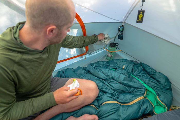 A man inside a tent sits on a Rabbit Ears 25 degree sleeping bag. A green water bottle and tent equipment are visible in the tent interior.