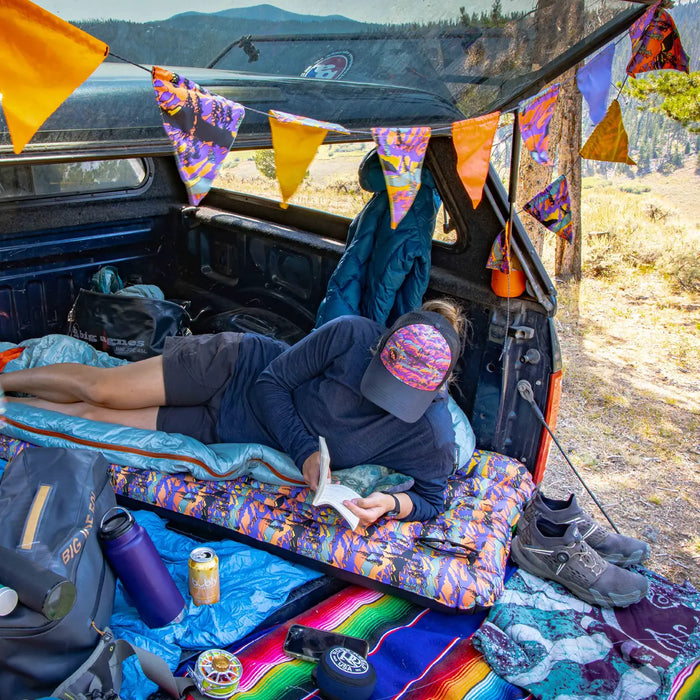 Relaxing in a pickup decorated with colorful flags, someone writes in a notebook while lying on the Big Agnes Rapide SL Insulated - Alpenglow sleeping pad. Camping gear, shoes, and drinks rest nearby against a stunning mountain backdrop.