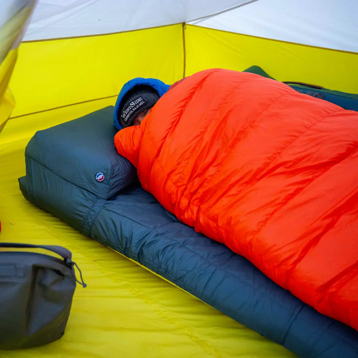 A person lies inside an orange sleeping bag on a Big Agnes Sleeping Pad Pillow Barn in a yellow tent, head covered and a bag in the foreground, enjoying extra comfort from the cozy camp pillow.