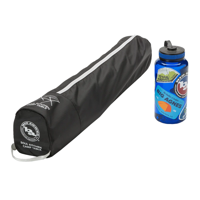 A black, cylindrical carrying case labeled Big Agnes Soul Kitchen Camp Table rests beside a blue sticker-covered water bottle, both set on a plain white background, ready for outdoor dining.