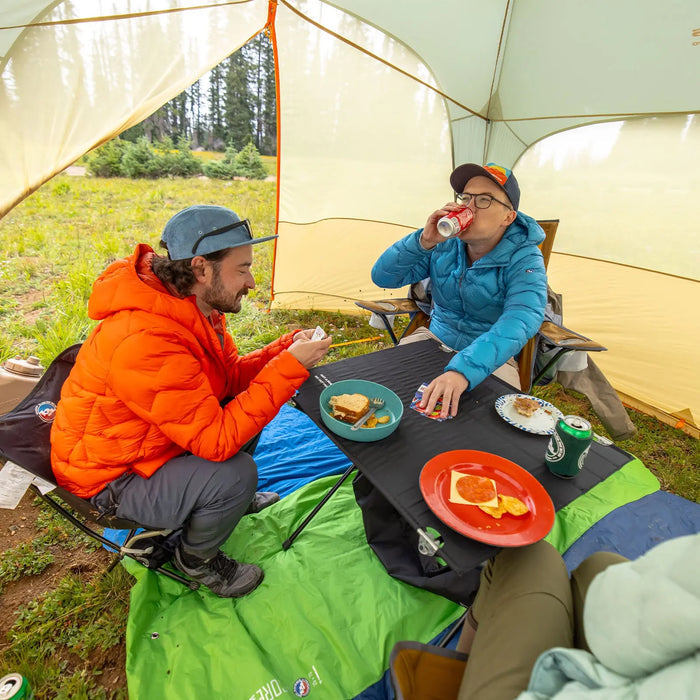 Two people in bright jackets relax under a tent at a campsite, enjoying meals and drinks on the Big Agnes Soul Kitchen Camp Table, which is covered with plates and snacks. Green grass and trees can be seen in the background.