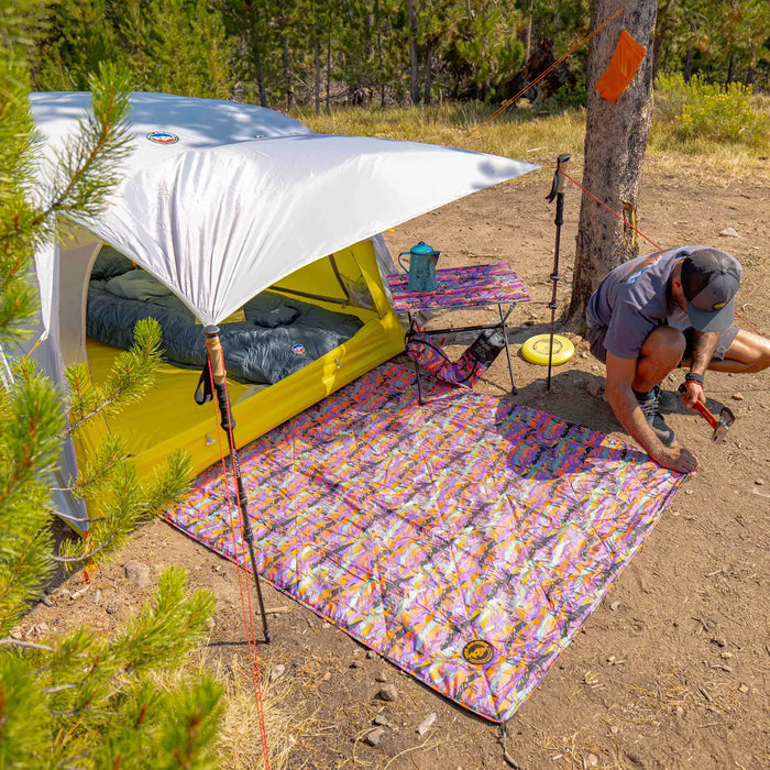 A person kneels by a vibrant Big Agnes Why Not Picnic Blanket - Alpenglow in front of a pitched tent at a forested campsite, with camping gear and chairs arranged nearby on a sunny day.