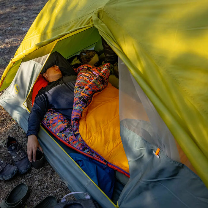 A person relaxes in a Big Agnes Women's Anthracite 20° - Alpenglow mummy sleeping bag inside a green tent, with camping gear and shoes outside, as sunlight filters through and casts gentle shadows across the campsite.