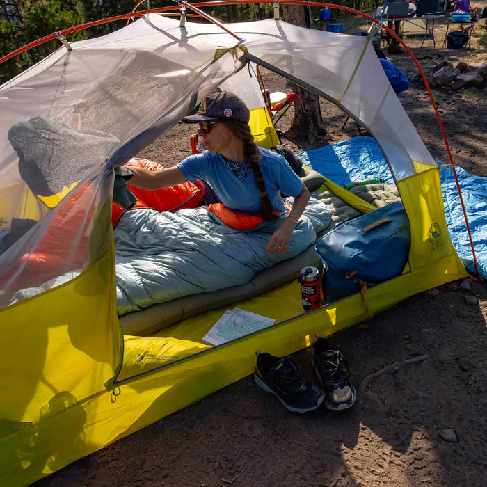 A camper wearing a cap sits in a yellow tent on a mat, reaching for the Big Agnes Women's Greystone 30° sleeping bag. Shoes, a map, and a blue backpack are outside. Trees and fellow campers can be seen in the background.