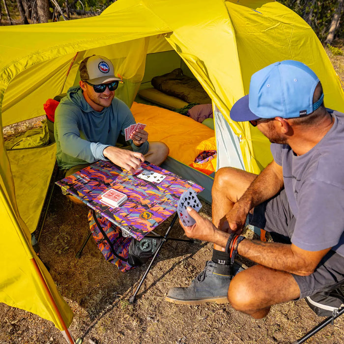 Two men sit outside a yellow tent at their campsite, playing cards on the Big Agnes Woodchuck Camp Table - Alpenglow. One deals cards while the other holds several, both clearly enjoying their game in the daylight.