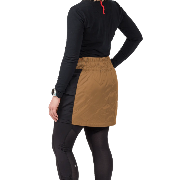 A person wearing black leggings, a black long-sleeve top, and the Big Agnes W's Columbine Skirt with diamond quilting stands with their back to the camera, also sporting a watch on their left wrist.