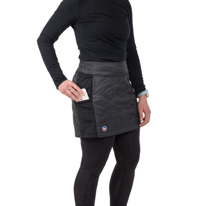 A woman wearing the Big Agnes W's Columbine Skirt in black with diamond quilting, paired with black leggings and a long-sleeve black top, places a smartphone in the skirt’s side pocket against a plain white background.