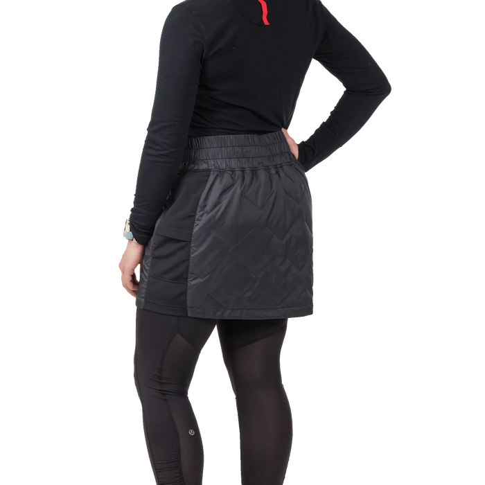 A person faces away from the camera, wearing a black long-sleeve top, black leggings, and the Big Agnes W's Columbine Skirt with diamond quilting, all set against a plain white background.