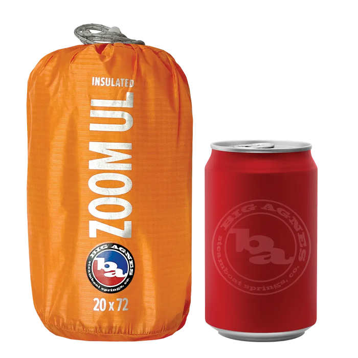 A compact, orange Big Agnes Zoom UL Insulated sleeping pad stands upright next to a red soda can, emphasizing its portable size. Both items are displayed against a plain white background.