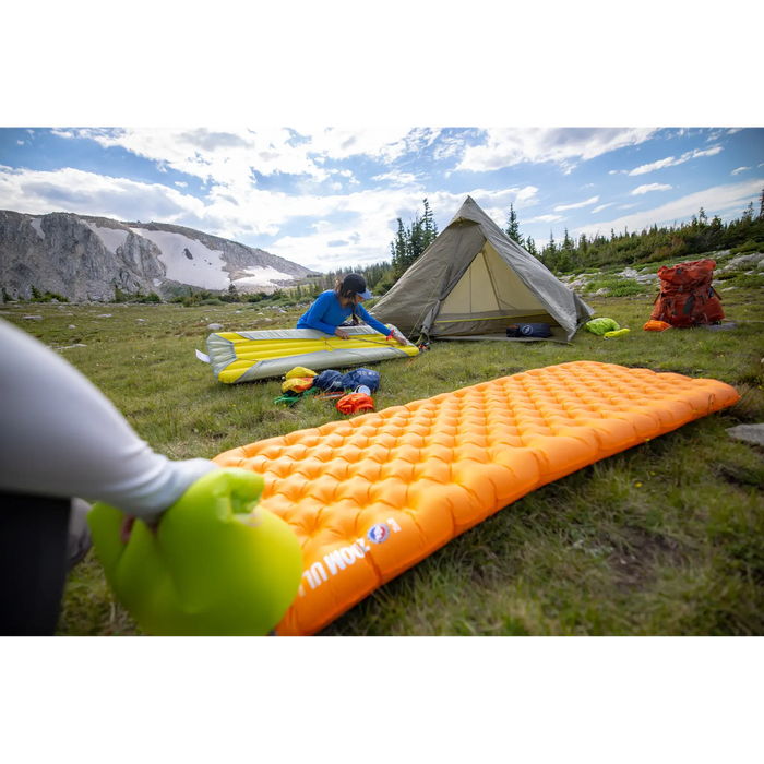 Two people set up camp in a grassy alpine meadow with mountain views. A tent is pitched nearby, and a Big Agnes Zoom UL Insulated sleeping pad and bright camping gear are scattered around the site.